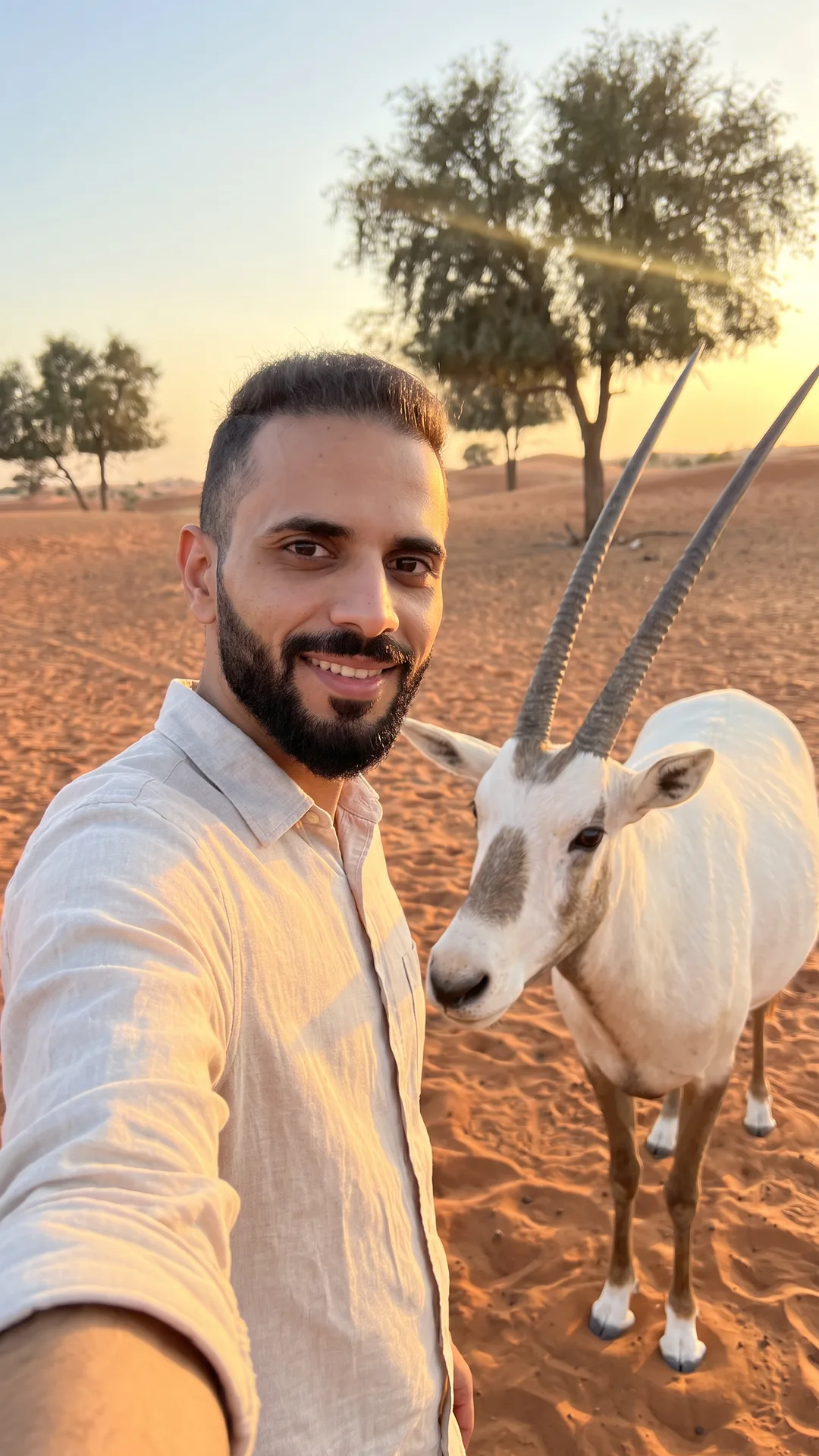 سيلفي مولّدة بالذكاء الاصطناعي لي بقميص كتان فاتح بجوار مها عربي أبيض طويل القرنين على رمال حمراء، وخلفنا أشجار غاف وضوء الغروب